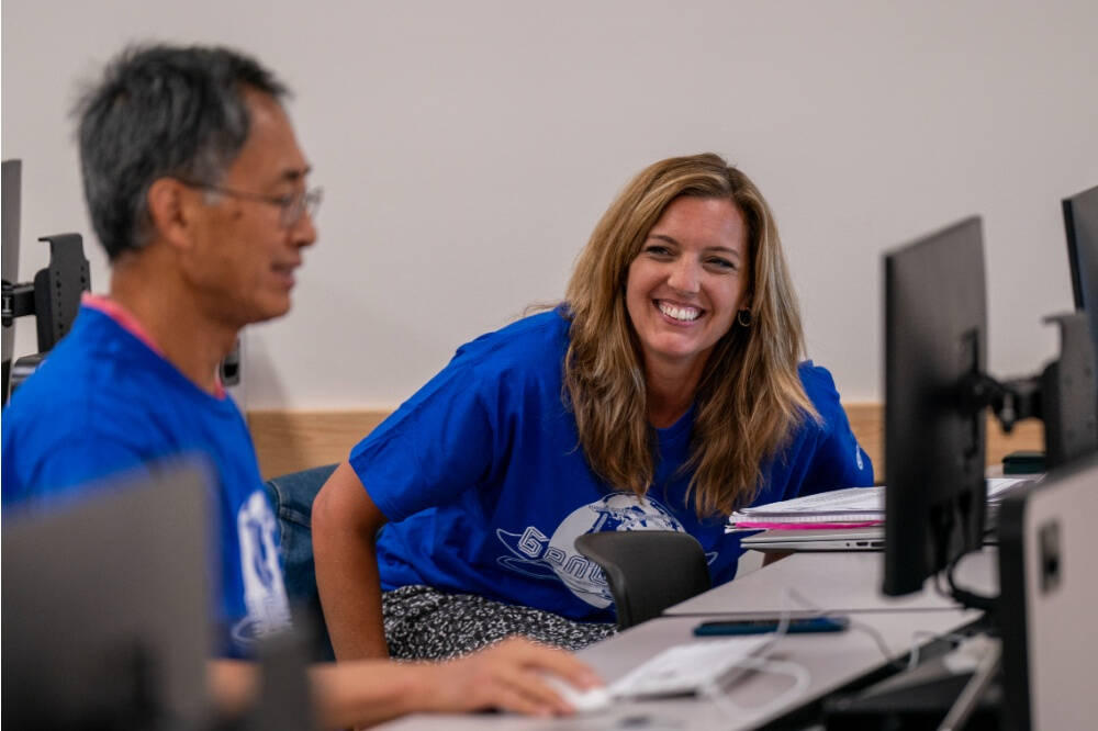 Two adults in blue T-shirts smile and interact in a computer lab, with the woman in focus laughing while seated next to computer monitors.